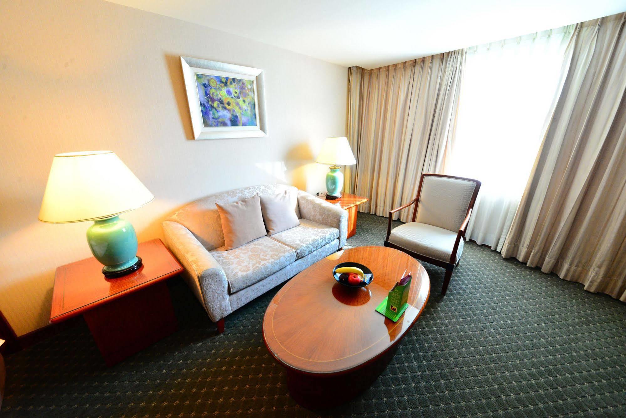 Evergreen Laurel Hotel - Taichung Exterior foto The photo shows a cozy living space, likely a hotel room or a lounge area. It features a light-colored sofa with cushions, a wooden coffee table in the center, and a comfortable armchair beside it. There are two lamps with turquoise bases on side tab