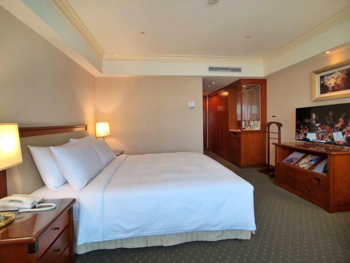 Evergreen Laurel Hotel - Taichung Exterior foto The photo shows a hotel room featuring a comfortable bed with white linens and pillows, positioned against a light-colored wall. There is a wooden bedside table with a lamp and an open book next to the bed. On the opposite side, there’s a large flat-