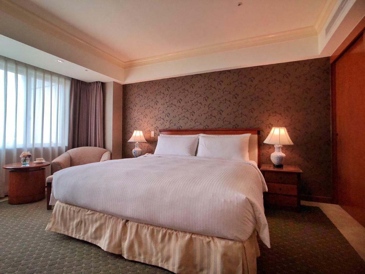 Evergreen Laurel Hotel - Taichung Exterior foto The photo shows a neatly arranged hotel room featuring a large bed with white linens and a light-colored bed skirt. On either side of the bed, there are small wooden nightstands with lamps. To the left of the bed, there's a comfortable chair and a ro