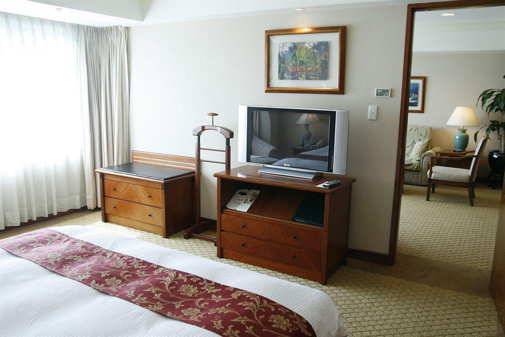 Evergreen Laurel Hotel - Taichung Exterior foto The photo depicts a hotel room interior. In the foreground, there is a neatly made bed with a decorative red bedspread. To the left, a large window allows natural light to fill the room, with light-colored curtains drawn to one side. 

Next to the be